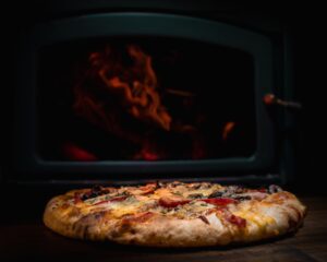 A baker using a long wooden peel to remove a blistered pizza from a glowing coal-fired brick oven.