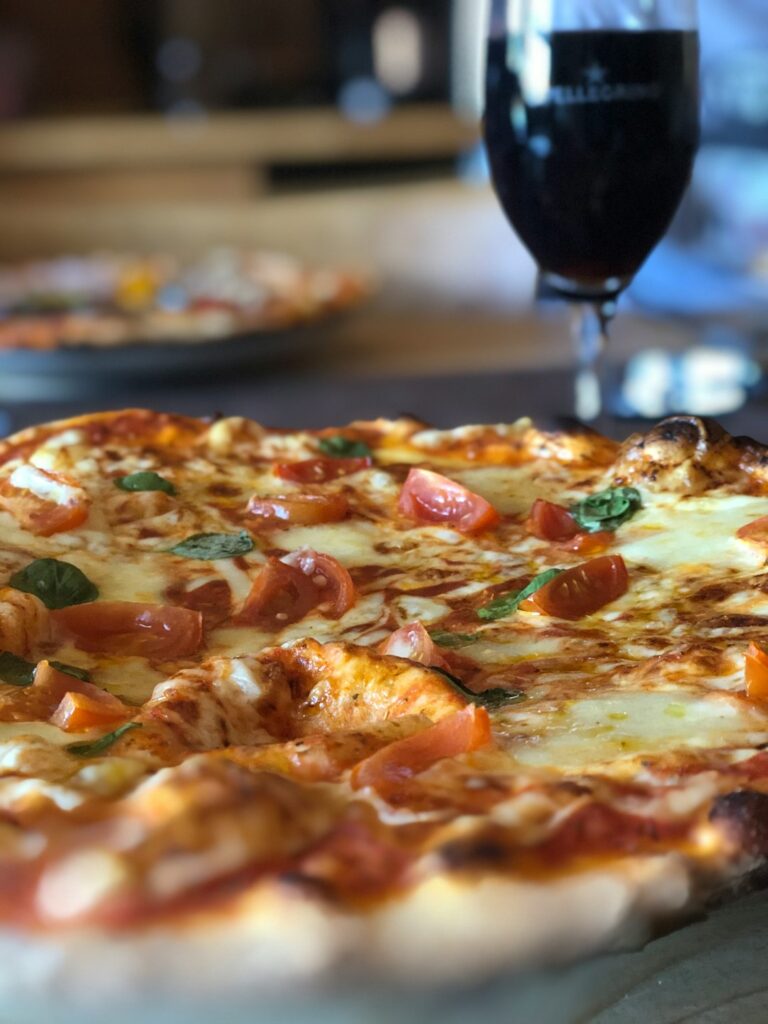 Two friends at a kitchen table enjoying pizza with glasses of wine and beer in a casual setting.