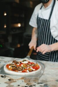 A person uses a pizza peel to place a homemade pizza into a kitchen oven with a visible pizza stone.