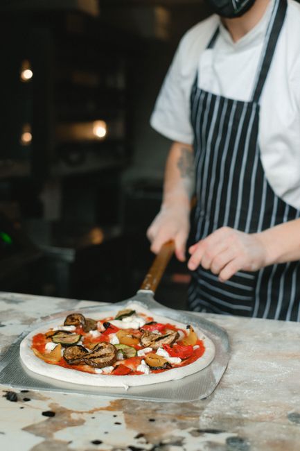 A person uses a pizza peel to place a homemade pizza into a kitchen oven with a visible pizza stone.